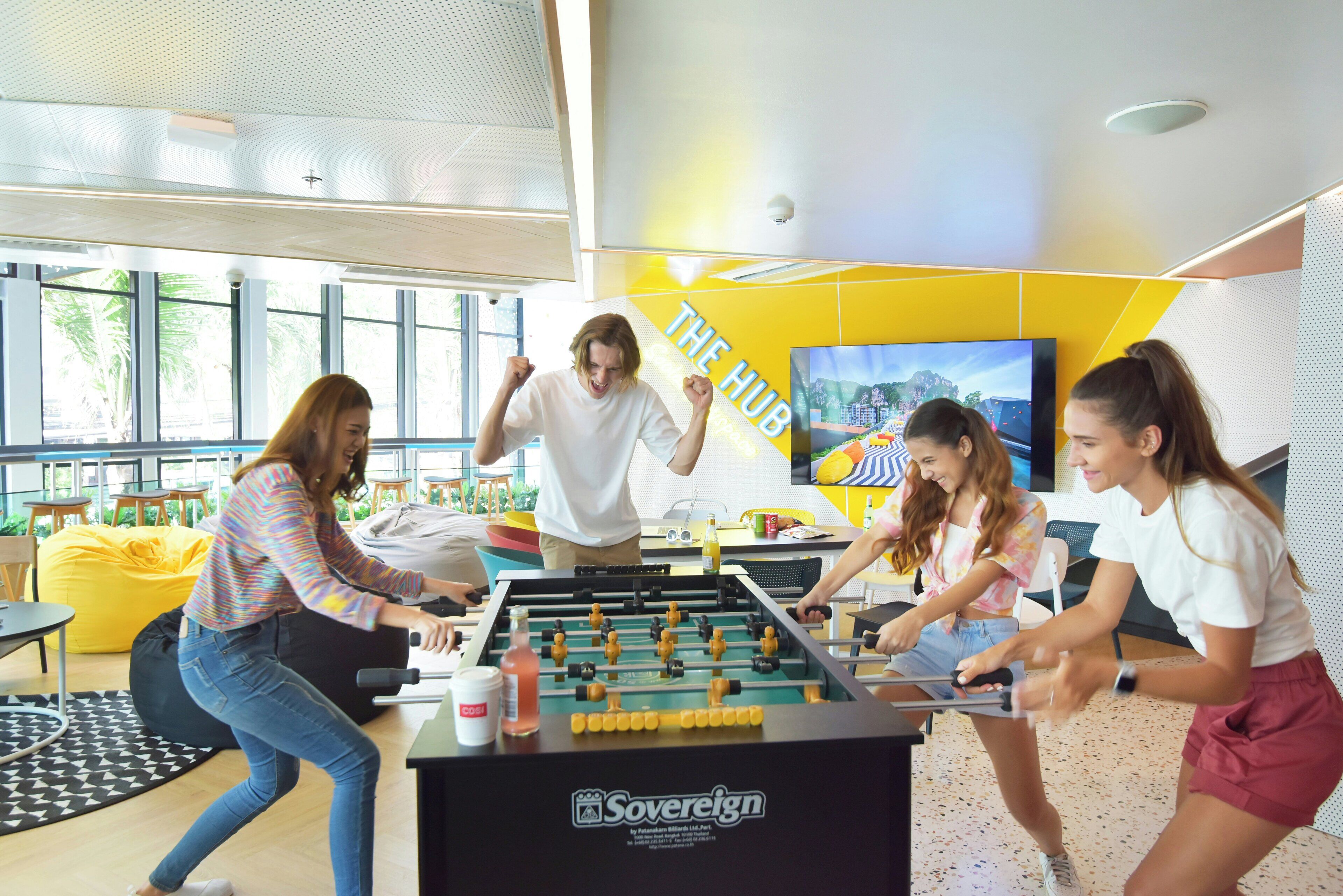 Un grupo de personas jugando al fútbol de mesa en un espacio interior luminoso con ventanas grandes y una pared amarilla.