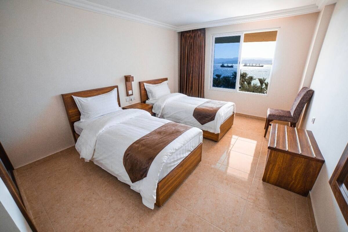 A hotel room with two beds, a wooden desk, and a view of the sea.