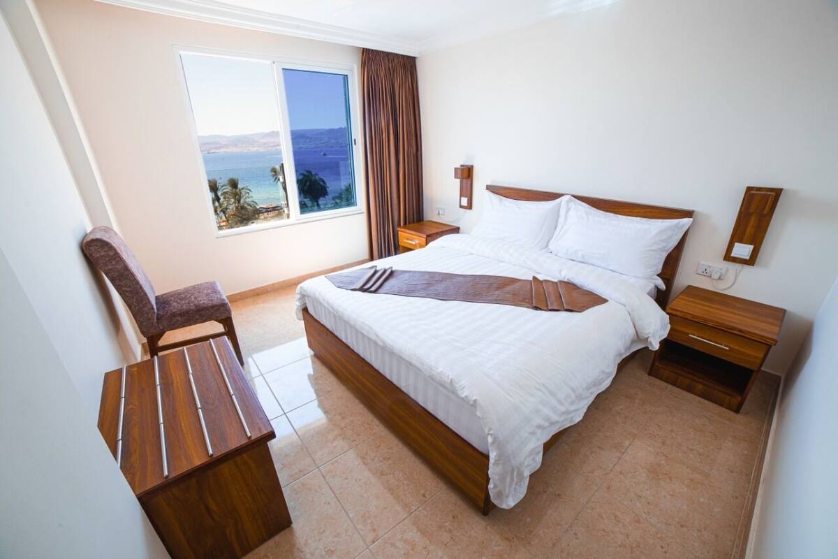 A hotel room with a bed, a wooden nightstand, a desk, a chair, a window with a view of the sea, and a curtain.