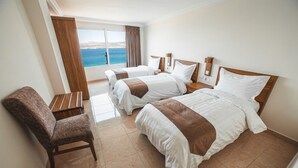 A hotel room with two beds, a chair, a nightstand, and a window with a view of the sea. - Joar Beach Hotel (Aqaba)