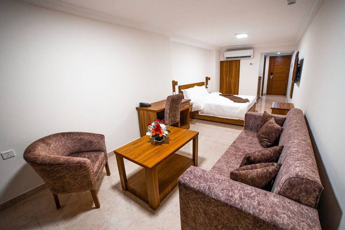 A modern hotel room with a bed, a wooden nightstand, a brown armchair, a sofa, and a small table with a vase.