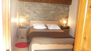 Een slaapkamer met een stenen muur, houten balken, een bed met witte beddengoed, twee bedlampen en een rood vloerkleed.