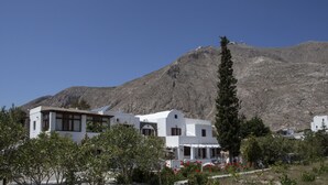 A white building with a flat roof and a large window, surrounded by greenery and a mountain in the background. - 3 Bedrooms Apartment included Car Rental (heated pool) (santorini)