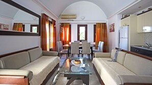 A modern living room with a glass coffee table, beige sofas, a dining area with chairs, and a kitchen with a refrigerator. - 3 Bedrooms Apartment included Car Rental (heated pool) (santorini)