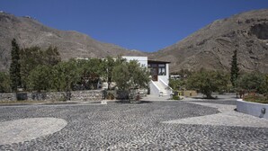 A modern building with a stone-paved courtyard, surrounded by trees and mountains. - 3 Bedrooms Apartment included Car Rental (heated pool) (santorini)