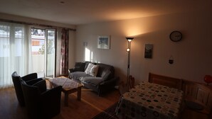Ein Wohnzimmer mit Sofa, Sesseln, Couchtisch, Esstisch mit gemustertem Tischläufer, Stehlampe, einer Uhr und einem Fenster mit Vorhängen.