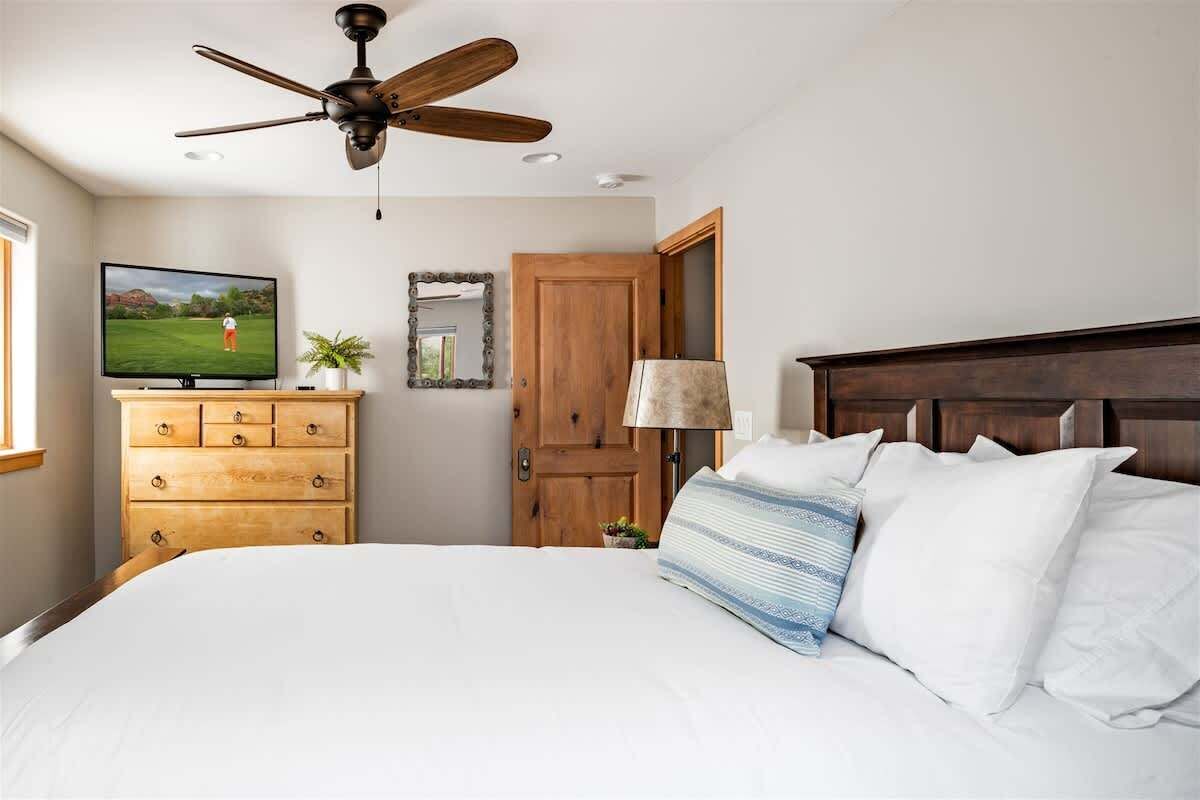 Quarto com cama, cômoda de madeira, ventilador de teto, televisão e uma porta.