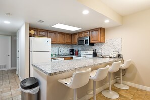 A kitchen with wooden cabinets, a stainless steel refrigerator, a microwave, and a granite countertop. There are two white bar stools and a trash can. - Sea Vista I #111 | Beachfront Ground-Floor Condo • Direct Beach Access • Two Community Pools (South Padre Island)