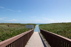A wooden boardwalk leading to a beach with a sign indicating no littering, no glass, no camping, leash and clean up after pets, and no shade devices between 9 AM - 9 PM.