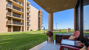 A covered outdoor patio with a barbecue grill, outdoor seating, and a view of a multi-story building.