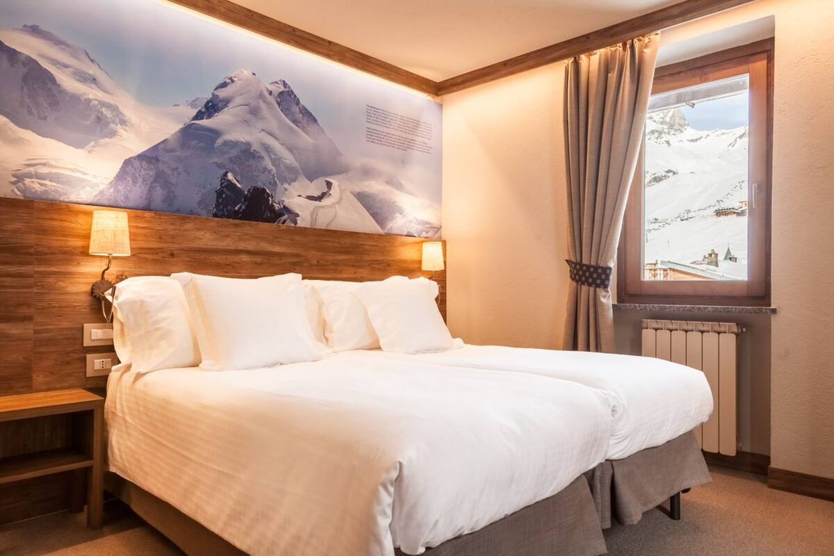 Habitación de hotel con una cama grande, cabecero de madera, dos lámparas de noche, una ventana con cortinas y un mural de montañas en la pared.