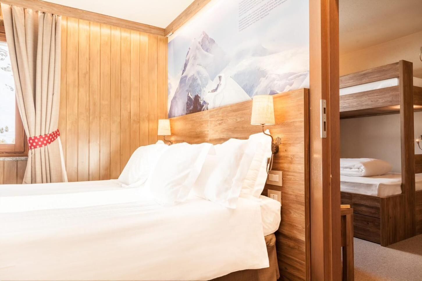 A hotel room with a wooden headboard, a bed with white linens, a window with beige curtains, and a mountain landscape painting on the wall.