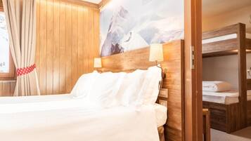 A hotel room with a wooden headboard, a bed with white linens, a window with beige curtains, and a mountain landscape painting on the wall.