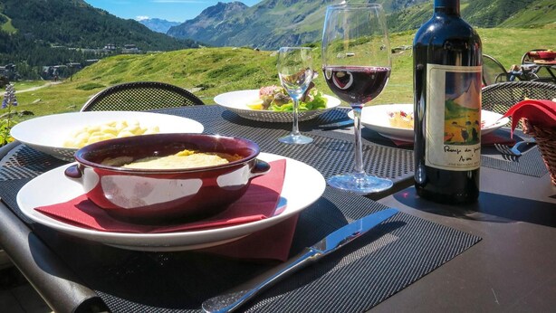 Une table dressée en plein air, avec une bouteille de vin, des verres et des assiettes remplies de nourriture, le tout sur fond de paysage montagneux.