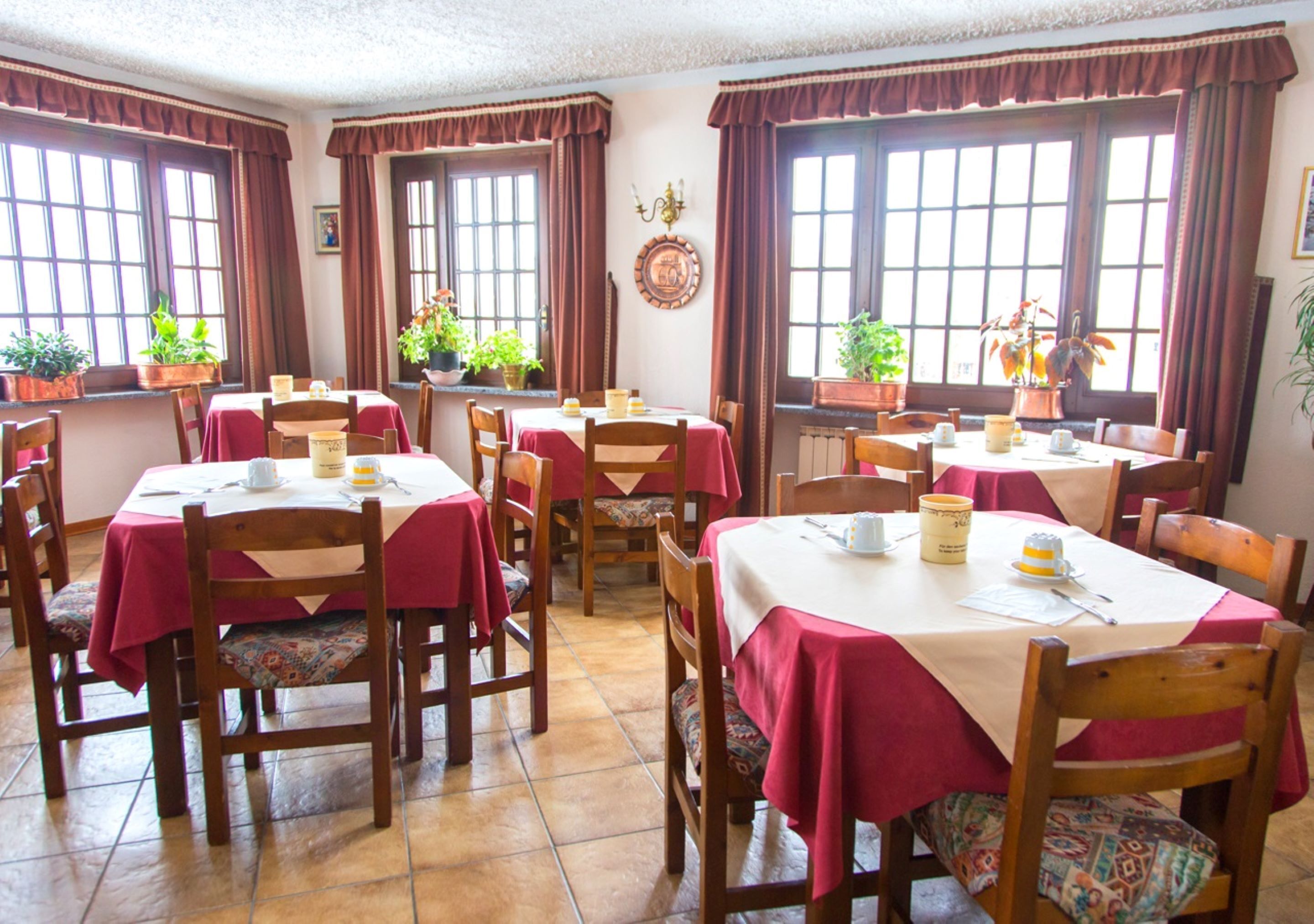 Zona pranzo con diversi tavoli e sedie, ognuno ricoperto da una tovaglia rossa. Sui davanzali ci sono piante in vaso e un orologio a parete.