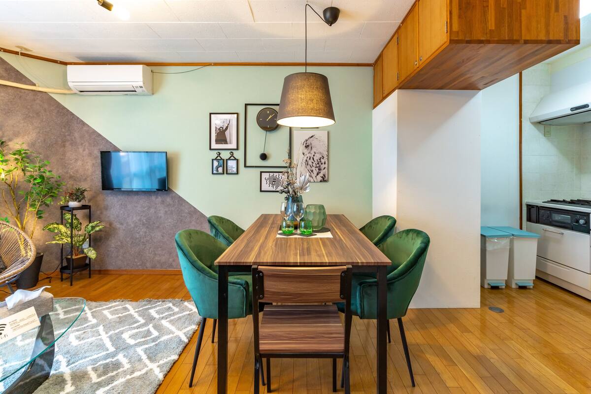 Un salón moderno con una mesa de comedor de madera, sillas verdes, un televisor montado en la pared y una cocina con armarios blancos y una estufa.