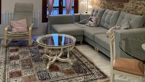 A living room with a grey sofa, a round glass coffee table, a beige armchair, a patterned rug, and a stone wall.