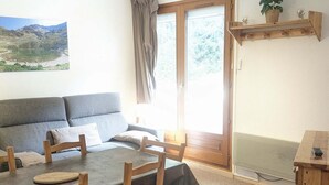 A room with a grey sofa, wooden dining table with chairs, a small wooden desk, and a window with a view of mountains. - 2-room cabin apartment at the foot of the slopes (Les Adrets-Prapoutel)