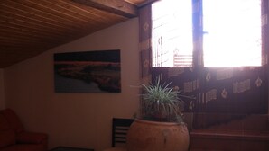 A dimly lit room with a wooden ceiling, a large window with a view of a staircase, a painting on the wall, a potted plant, a sofa, and a small table.