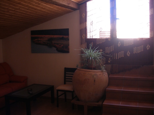 A dimly lit room with a wooden ceiling, a large window with a view of a staircase, a painting on the wall, a potted plant, a sofa, and a small table.