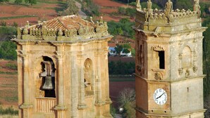 A historic tower with a bell and a clock, surrounded by terraced fields and a winding road.