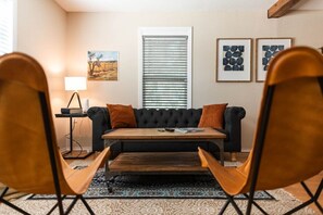 Ein Wohnzimmer mit einer schwarzen Couch, einem Holztisch und zwei Holzstühlen. Ein Fenster mit Jalousien, eine Lampe und gerahmte Kunstwerke sind an der Wand zu sehen.