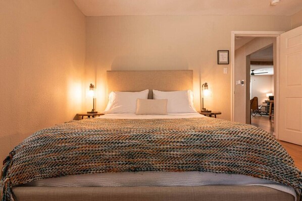 Ein Hotelzimmer mit einem Bett, zwei Nachttischlampen, einer Tür und einem Gang zu einem anderen Zimmer.