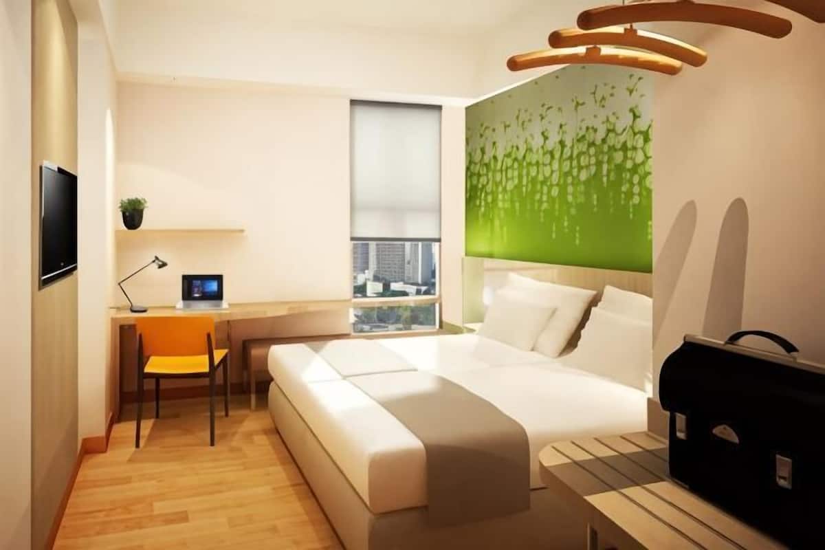 Moderni hotellihuone, jossa on sänky, työpöytä tietokoneella, tuoli, televisio ja ikkuna, josta avautuu näkymä kaupunkiin.