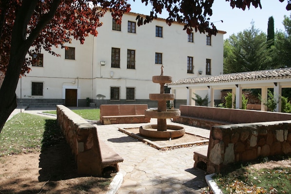 Un patio con una fuente de piedra, bancos y un edificio blanco con múltiples ventanas al fondo.