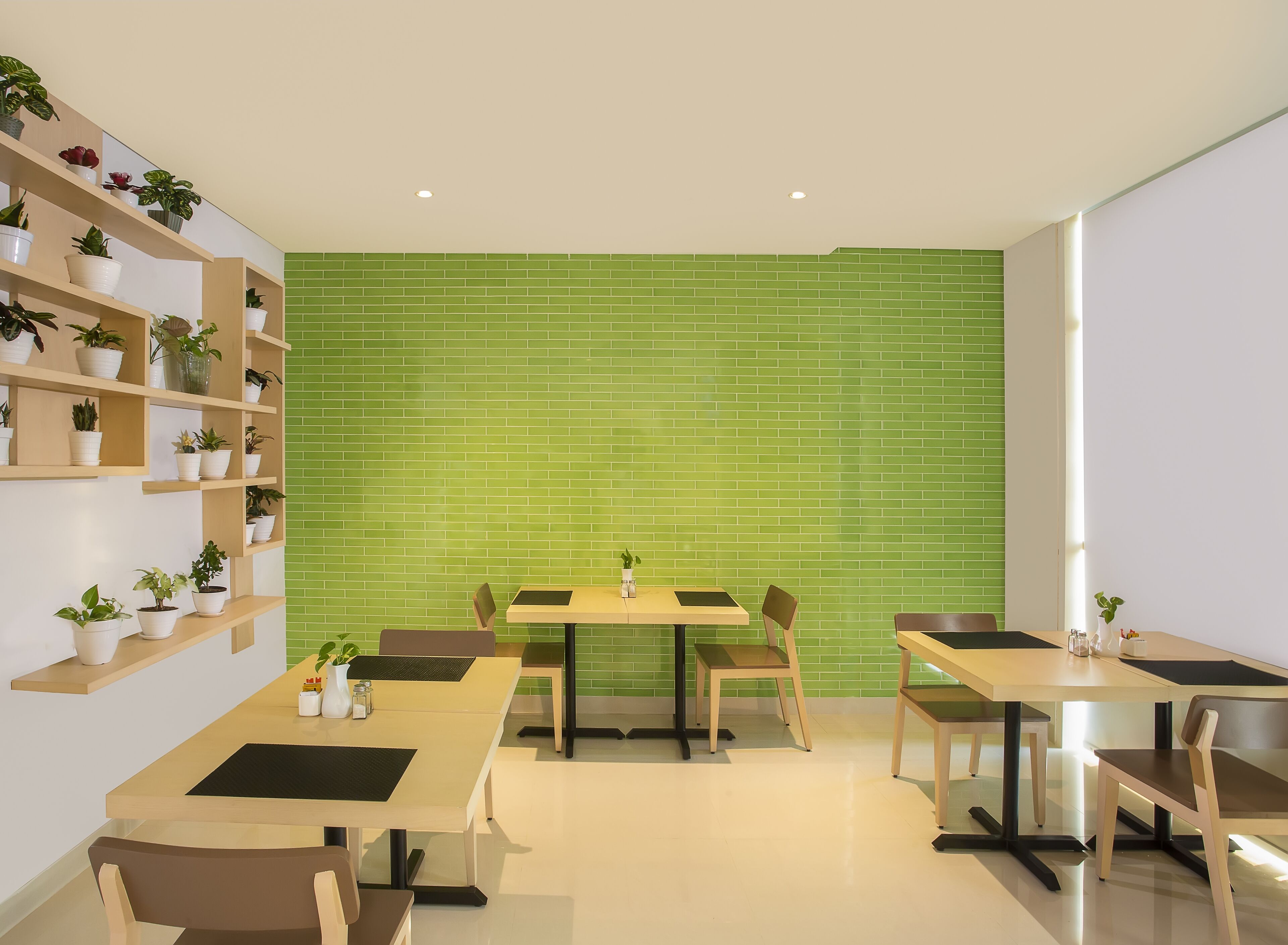 A modern dining area with green-tiled walls, wooden tables, and chairs, and shelves with potted plants.