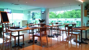 Un espace de restauration moderne, avec des tables et des chaises, de grandes fenêtres donnant sur un paysage verdoyant et un téléviseur fixé au mur.