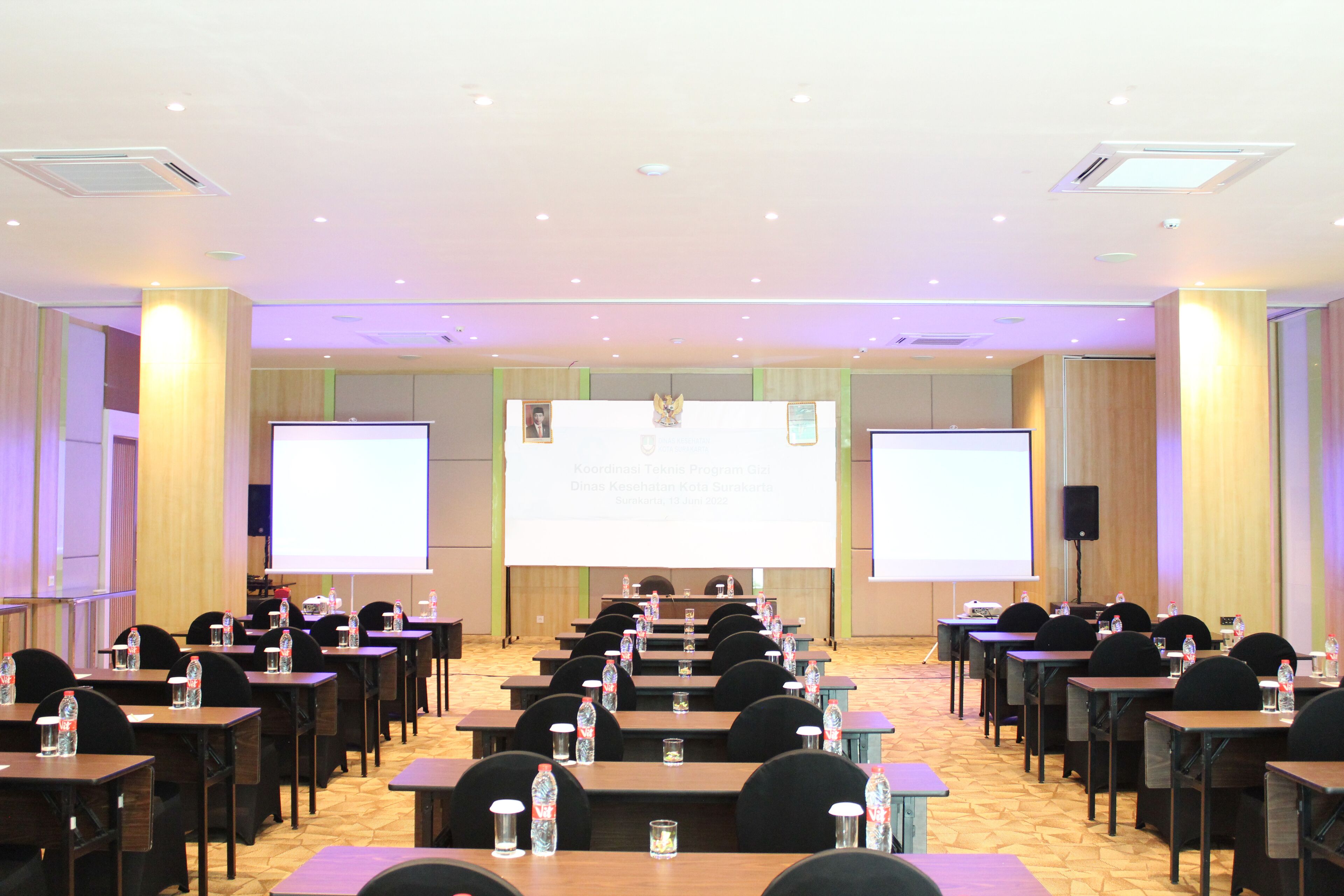 Un salón de conferencias con filas de mesas y sillas, equipado con proyectores y pantallas, y botellas de agua en cada mesa.