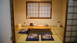 Une pièce de style japonais traditionnel, avec un sol recouvert de tatamis, un paravent en papier de riz (shoji), une petite table sur laquelle sont posés des livres et une petite lampe.