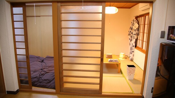 Une pièce de style japonais traditionnel, avec une porte coulissante, un lit recouvert d’une couette violette, une petite table avec une lampe et un téléviseur posé sur un meuble.