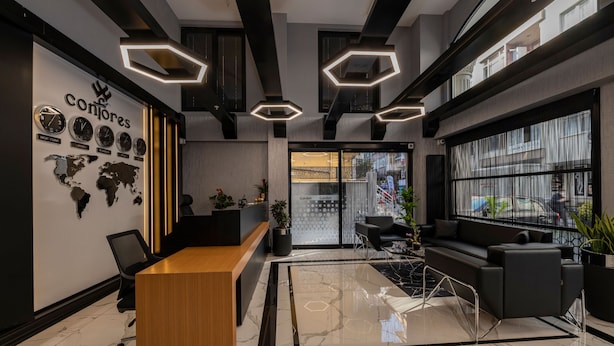A modern reception area with a wooden reception desk, black sofas, and a large window offering a view of the street. - Confores Hotel (Istanbul)