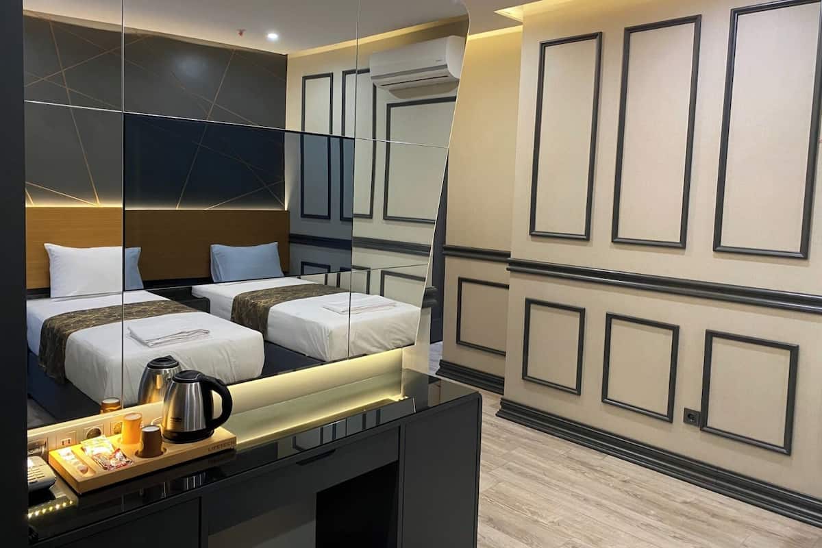 A modern hotel room with a large mirror, two beds, a desk with a coffee maker, and a minibar.