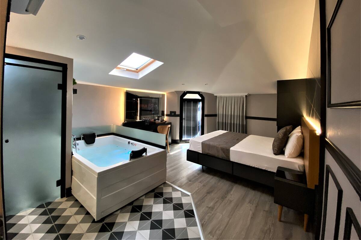 A modern hotel room with a skylight, a separate bathroom, a large bathtub, and a bed with a nightstand.