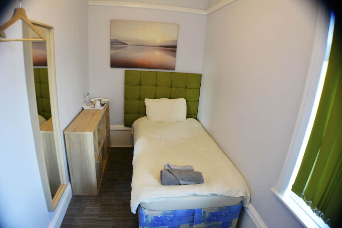 Una habitación de hotel pequeña y ordenada con una cama individual, una mesita de noche con lámpara, un espejo y un cuadro en la pared.