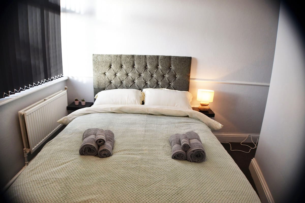 Una cama bien hecha con tablero de cabecera capitonado, dos almohadas y toallas enrolladas sobre la cama.