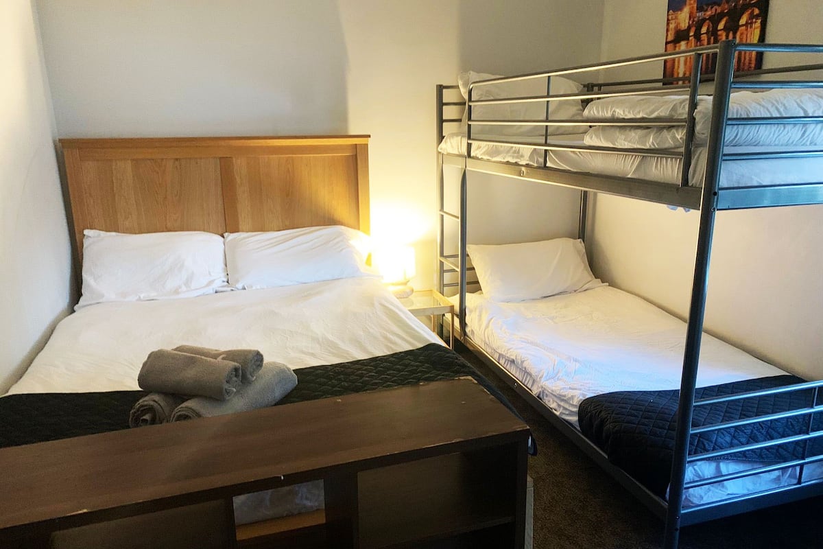 Habitación de hotel con una cama individual, cabecera de madera, mesita de noche con lámpara y una cama litera de dos niveles.
