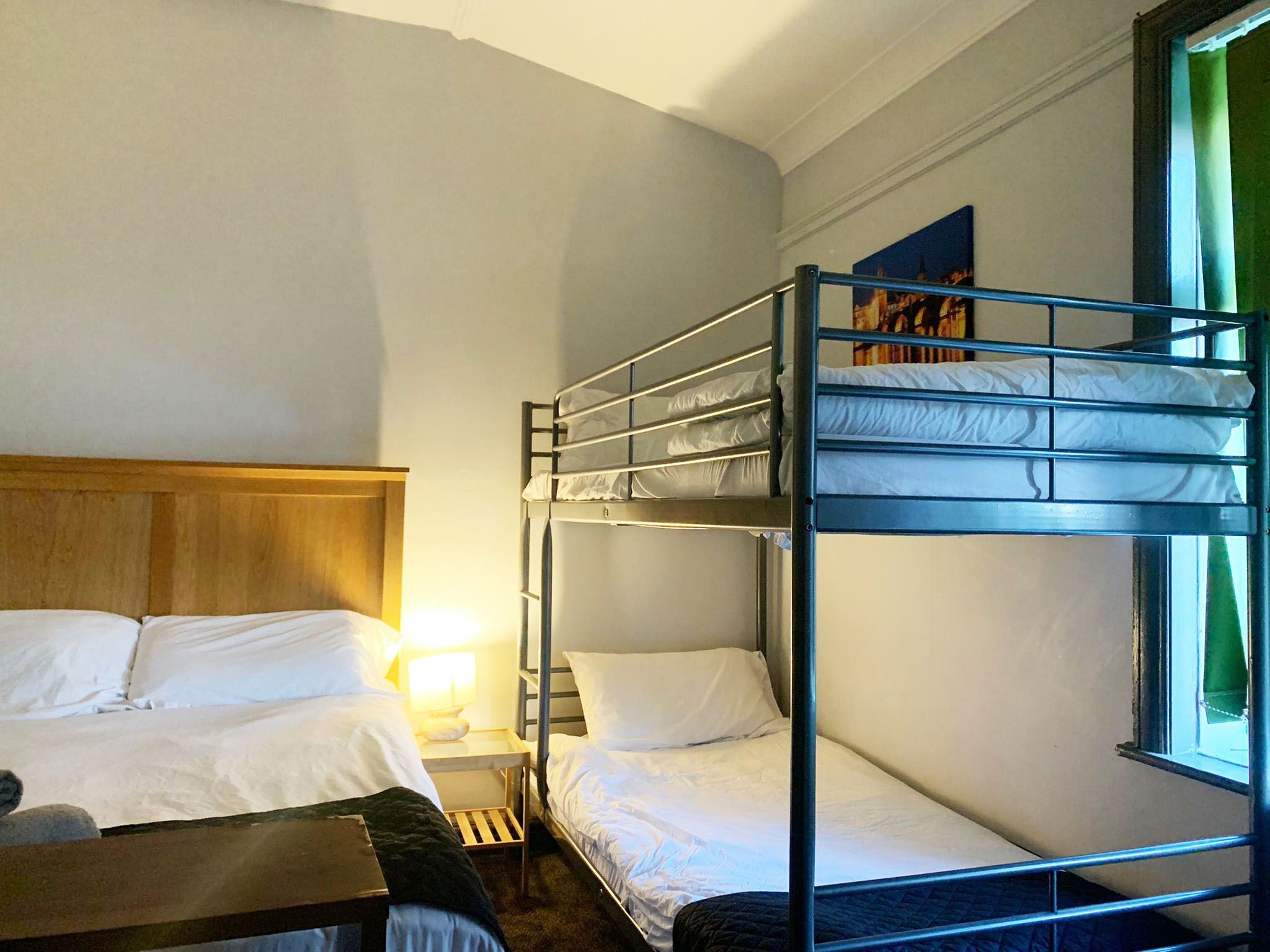 Dormitorio con literas, cabecera de madera, una cama individual, mesita de noche con lámpara y ventana con cortinas.