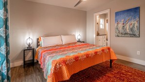 Un dormitorio con cama, ventilador de techo, mesita de noche, un cuadro y baño visible a través de una puerta abierta.