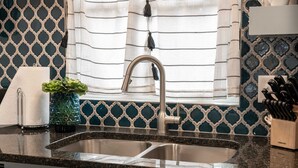 Una cocina con un lavamanos de acero inoxidable, una ventana con cortinas, una planta en maceta y un bloque de cuchillos.