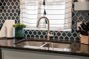 Una cocina con un lavamanos de acero inoxidable, una ventana con cortinas, una planta en maceta y un bloque de cuchillos.