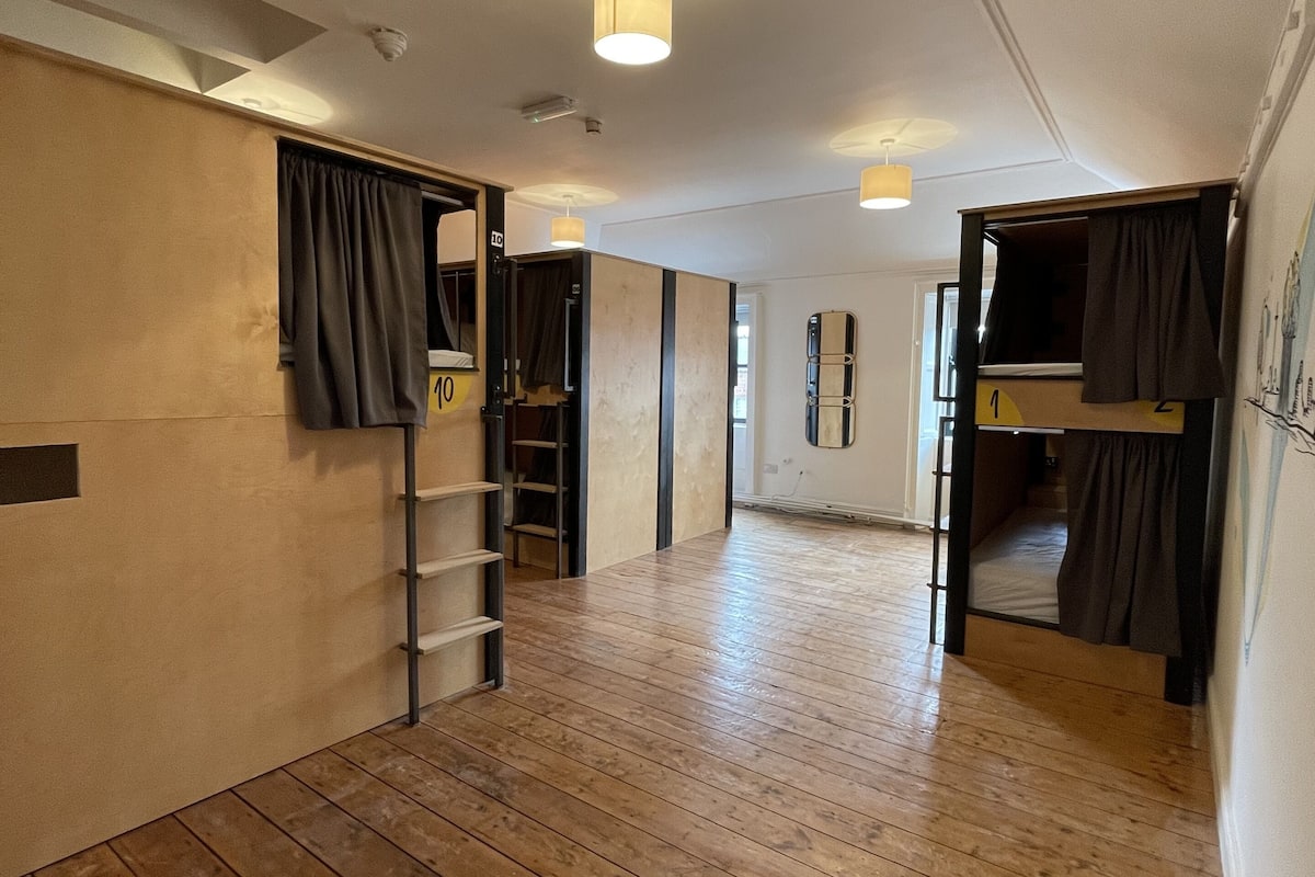 Una habitación amplia con suelo de madera, una cama litera, una escalera y un espejo en la pared.