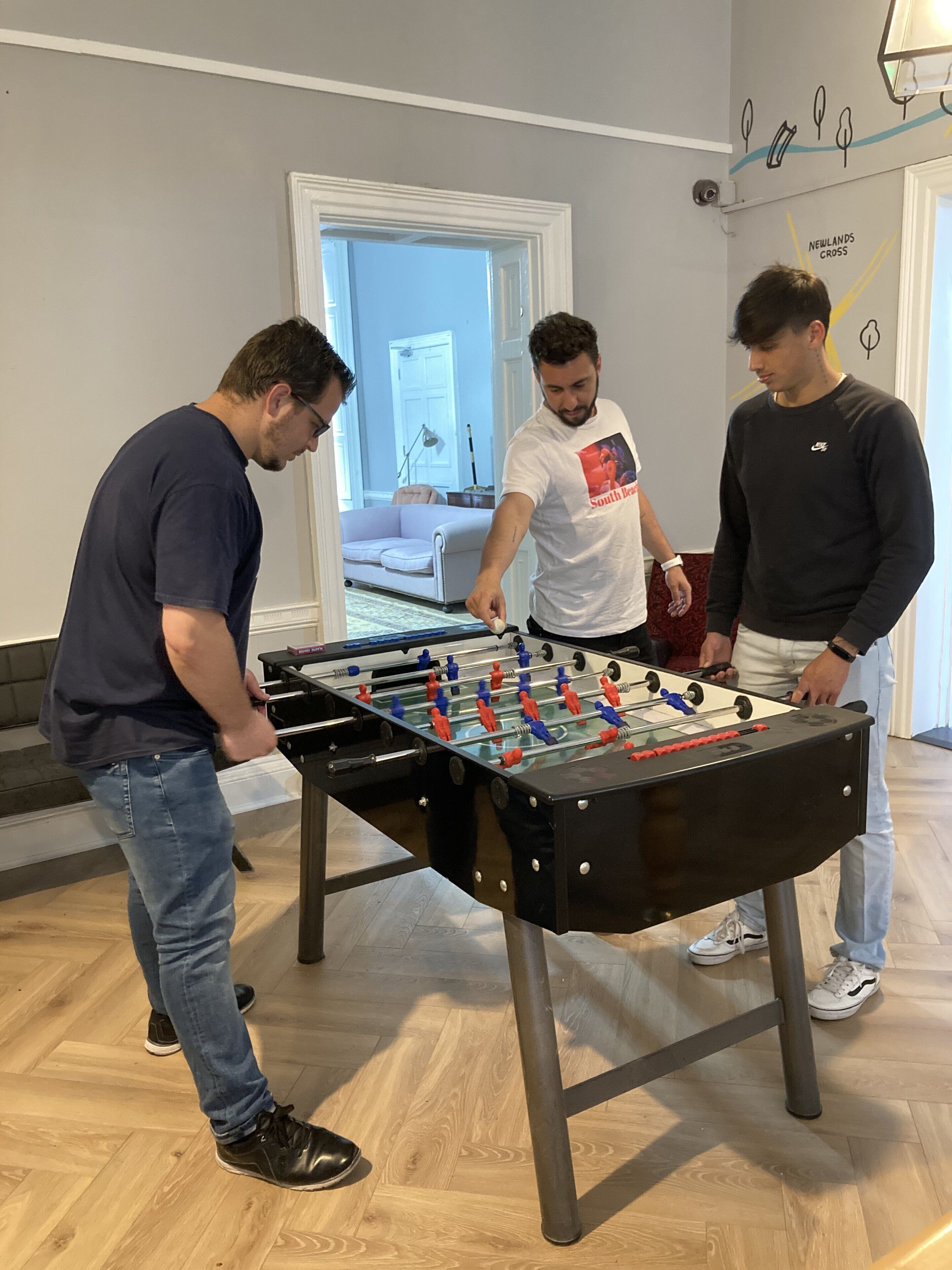A group of people playing foosball in a room with a wooden floor and a white sofa in the background.
