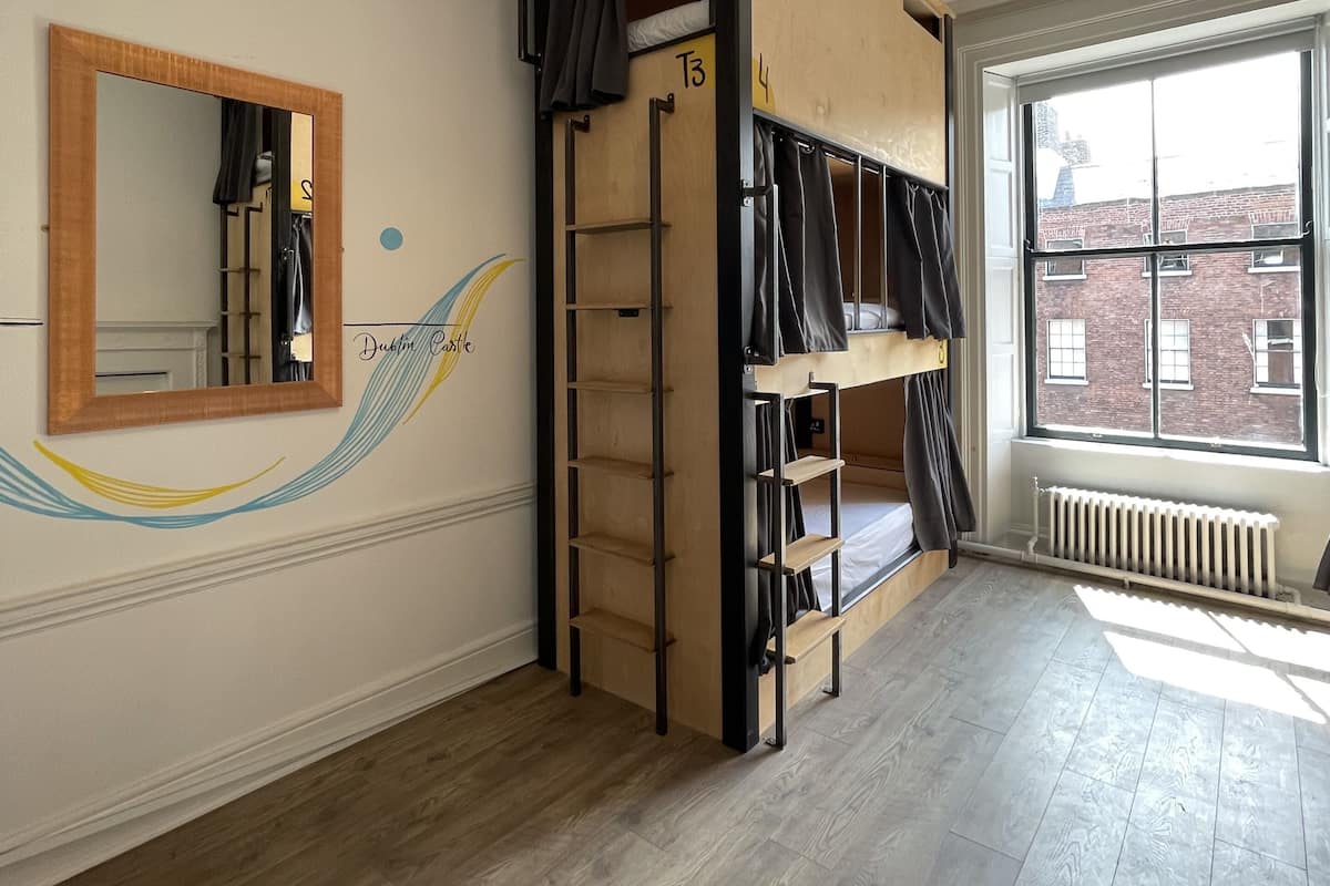 Una habitación con literas, un espejo, un radiador y una ventana que ofrece vistas a un edificio de ladrillo.