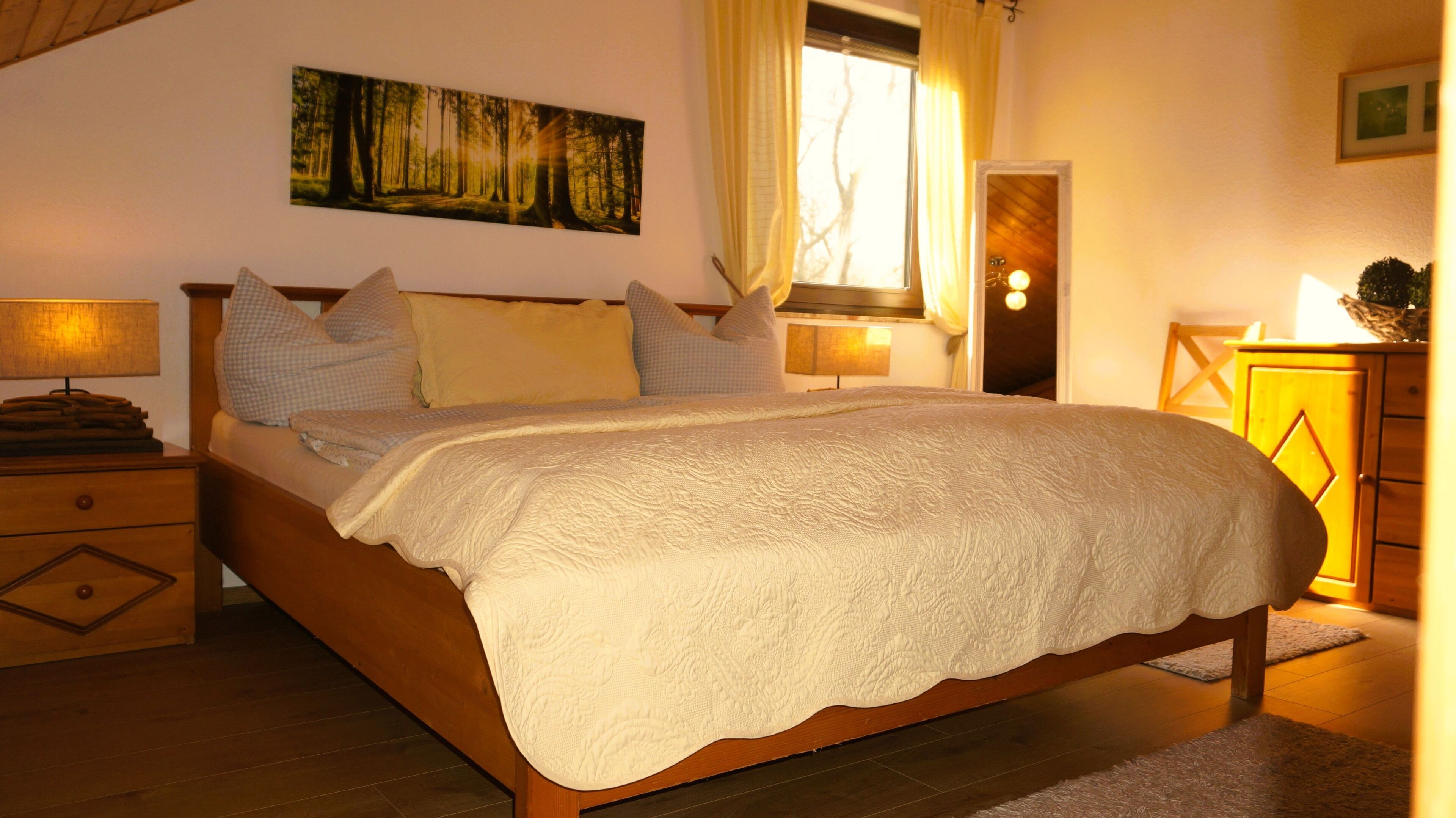 A bedroom with a large bed, a painting of a forest, a window with curtains, and a wooden cabinet.