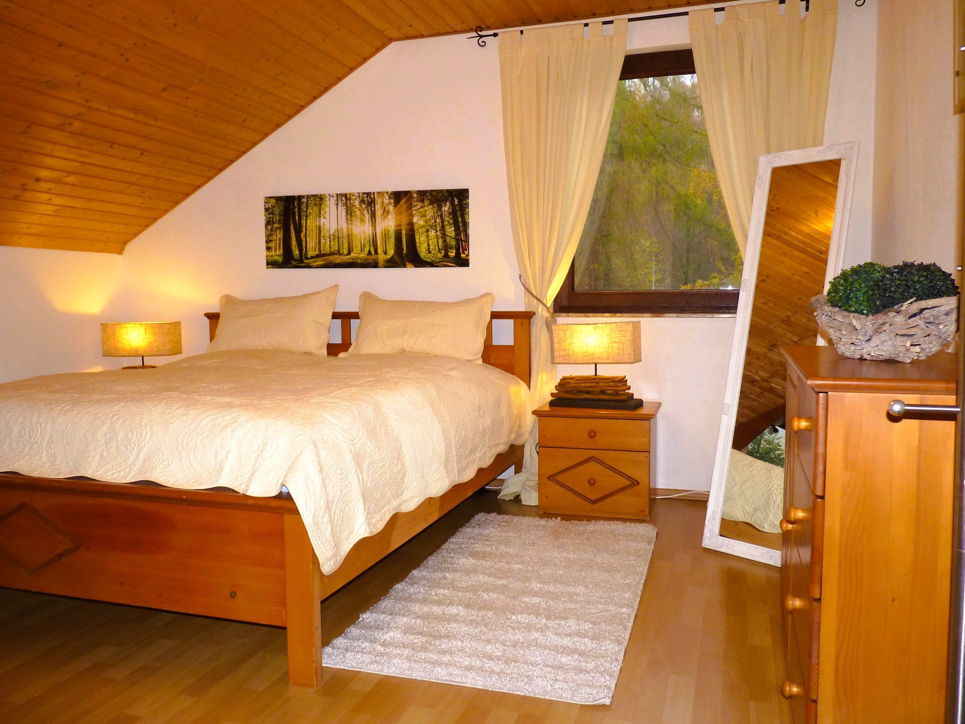 A bedroom with a wooden bed, a nightstand with a lamp, a dresser, a window with curtains, and a painting on the wall.