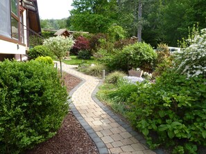 A garden pathway leading to a wooden structure, surrounded by lush greenery and various plants.